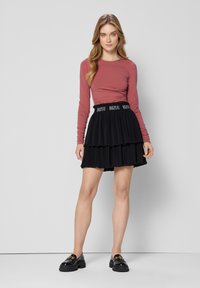 Langsleeve, strakke, rozenkleurige top gecombineerd met een zwarte, gelaagde rok met een logo tailleband. Schoenen zijn zwarte instappers met gouden hardware.