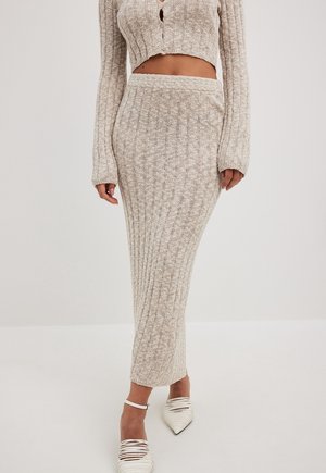 Woman wearing a beige ribbed knit long skirt, cropped long-sleeve top, and white strappy pointed-toe shoes standing on light background.
