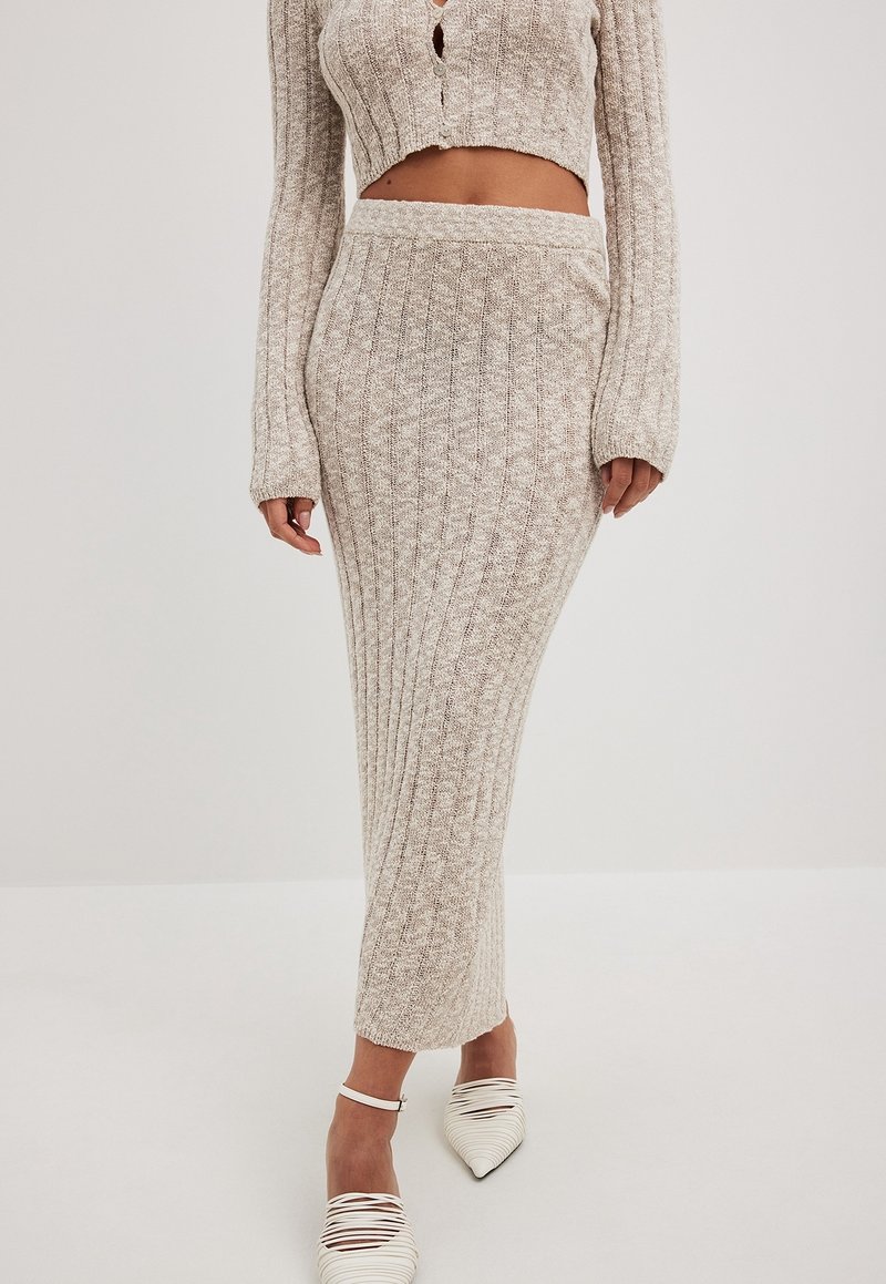 Femme portant une longue jupe en tricot côtelé beige, un top court à manches longues et des chaussures blanches à brides et bouts pointus, debout sur un fond clair.