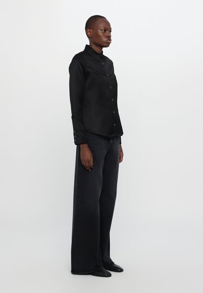 Svart knapp-up skjorte med krage, innsnevret i midjen, paret med brede mørkeblå denimbukser. Glatt tekstur, minimalistisk design.
