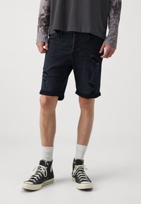 Pantalones cortos de mezclilla oscura con un diseño desgastado, dobladillos enrollados y rasgaduras visibles. Combinados con zapatillas negras y calcetines blancos hasta la pantorrilla.