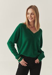 Grüner Pullover mit V-Ausschnitt, lockerer Passform und langen Ärmeln. Getragen über schwarzen Hosen mit gerippten Bündchen und glatter Textur.