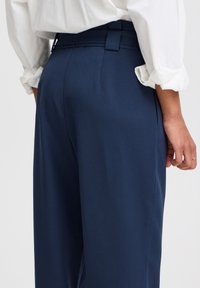 Pantalons taille haute bleu marine avec une coupe ajustée, présentant une texture lisse et des coutures, complétés par des passants de ceinture et un élégant tailoring.