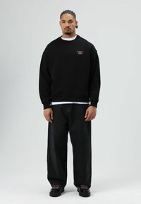 Zwarte sweatshirt met geribbelde manchetten, gouden logodetail, gecombineerd met losvallende zwarte jeans en chunky zwarte schoenen op een effen achtergrond.