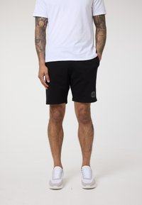 Juodi medvilniniai šortai su elastinga juosta, šoninėmis kišenėmis ir tekstūruotu apvaliu logotipu kairėje šlaunyje; derinami su baltais sportbačiais.