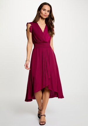 Kvinde iført ærmeløs dyb pink kjole med høj-lav kant og flæseærmer samt sorte hæle med ankelrem, stående foran en ensfarvet baggrund.