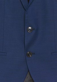 Gros plan sur un blazer bleu texturé avec deux boutons noirs fermés, montrant les poches avant et une partie de la doublure intérieure noire.