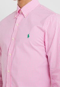 Růžová kostkovaná košile na knoflíky s klasickým límcem, jemnou texturou a malým zeleným vyšitým logem na hrudi.