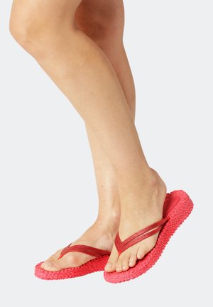 Barfuß mit roten strukturierten Flip-Flops, ein Fuß leicht angehoben, vor einem einfarbigen hellgrauen Hintergrund.