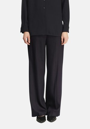 Kobieta ma na sobie czarną koszulę na guziki oraz szerokie czarne spodnie z czarnymi, spiczastymi butami, stoi na białym tle.