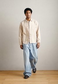 Beżowa bawełniana kurtka z dwoma kieszeniami na piersi, zestawiona z jasnoniebieskimi szerokimi jeansami i ciemnobrązowymi butami, noszona przez osobę idącą.