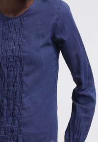 Mörkblå blus med långa ärmar och strukturerad vertikal veckad design på framsidan, bärs av en person mot en neutral bakgrund.