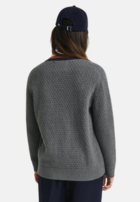 Grått texturerat tröja med lös passform, med ribbad fåll och krage. Långa ärmar med ett vävt mönster, bärs med en mörk keps.