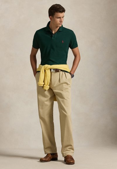Ung mand iført en mørkegrøn polo, beige bukser, brune sko og en gul sweater bundet om livet, stående med hænderne i lommerne.