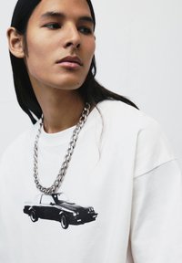 Hvid sweatshirt med et sort bilgrafik på forsiden. Modellen bærer en kraftig sølvkæde. Glat stof med løs pasform.