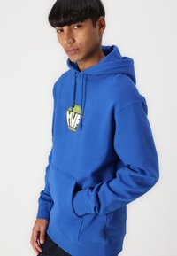 Blue hoodie in cotton, featuring a front pocket and a logo patch. It has a drawstring hood and ribbed cuffs, showcasing a soft texture.