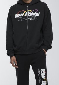 Sudadera negra con capucha y cremallera, con texto colorido "low lights" en el pecho, y pantalones deportivos negros a juego con logo y texto en la pierna.