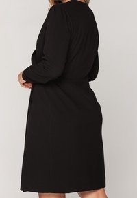 Svart långärmad klänning med en figurnära silhuett, bälte i midjan och mjuk textur. Har en enkel design och medellång längd.
