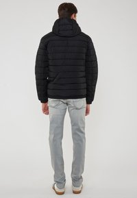 Piumino nero con cappuccio, design trapuntato e cuciture verticali, abbinato a jeans grigi chiari e scarpe casual.