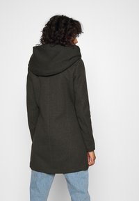 Cappotto verde scuro con un ampio cappuccio piegato, vestibilità su misura e una consistenza delicata, abbinato a jeans azzurri chiari.