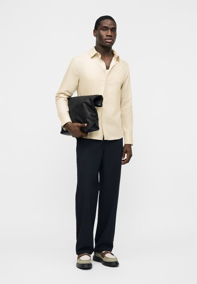 Άνδρας που φοράει μπεζ πουκάμισο και σκούρο παντελόνι κρατά διπλωμένη μεγάλη μαύρη δερμάτινη θήκη, στέκεται μπροστά σε απλό λευκό φόντο.