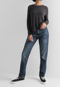 Top lungo grigio scuro a maniche lunghe, vestibilità morbida, abbinato a jeans blu a gamba dritta e sneakers nere, in piedi su uno sfondo grigio neutro.