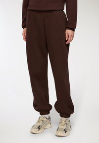 Brązowe dresy wykonane z miękkiego, elastycznego materiału. Posiadają elastyczny pas i ściągacze przy kostkach. Na nogach są beżowe sneakersy.