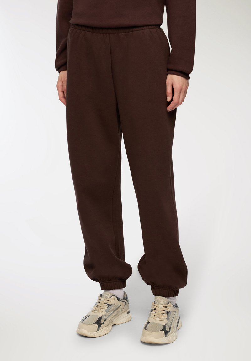 Brązowe dresy wykonane z miękkiego, elastycznego materiału. Posiadają elastyczny pas i ściągacze przy kostkach. Na nogach są beżowe sneakersy.