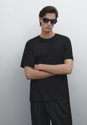 Black cotton t-shirt with short sleeves, crew neck; paired with black trousers. Accessories include rectangular black sunglasses.