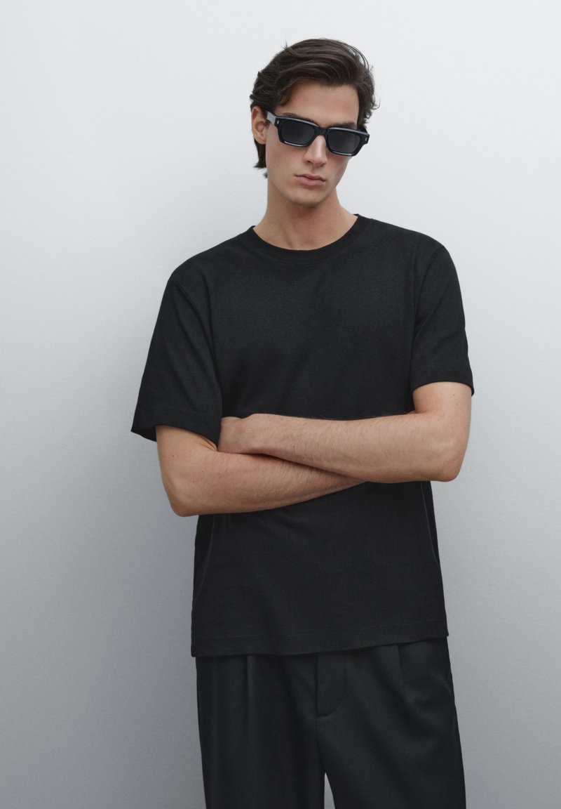 Schwarzes Baumwoll-T-Shirt mit kurzen Ärmeln und Rundhalsausschnitt; kombiniert mit schwarzen Hosen. Die Accessoires umfassen rechteckige schwarze Sonnenbrillen.