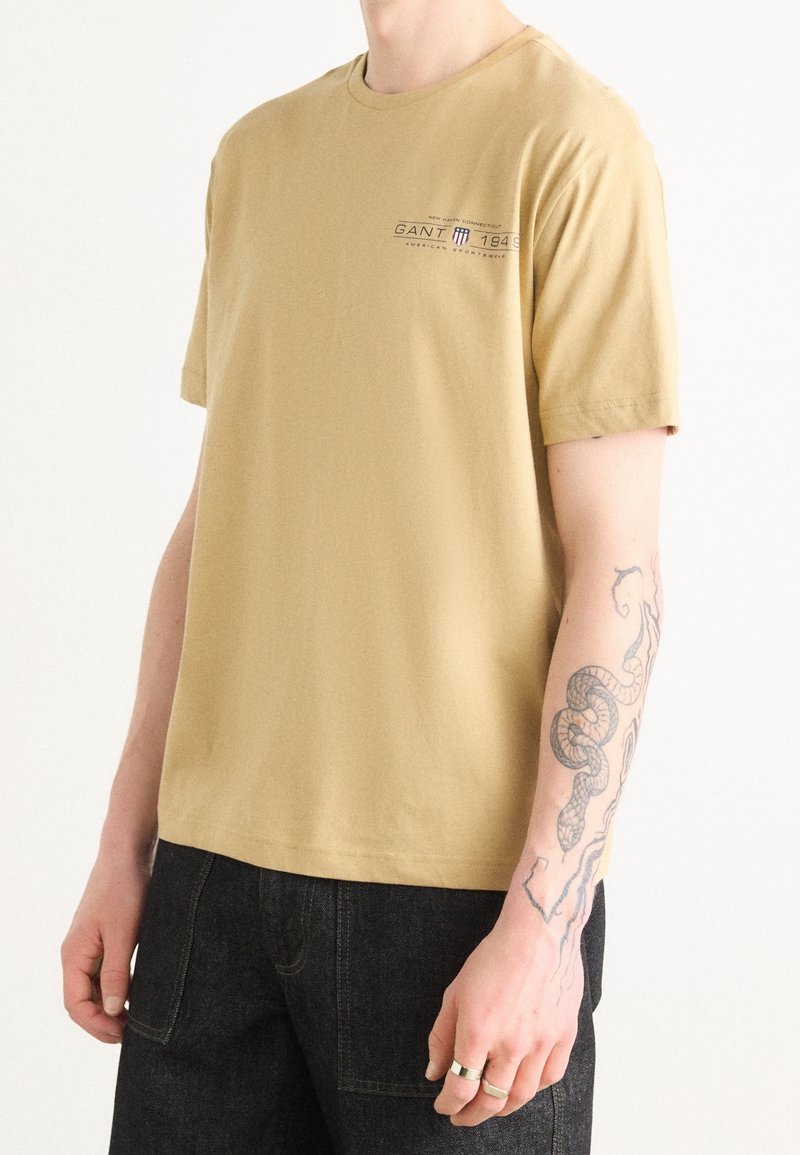 T-shirt à manches courtes, col rond en coton beige. Présente un petit logo imprimé sur la poitrine et une coupe décontractée. Associé à un jean en denim foncé.