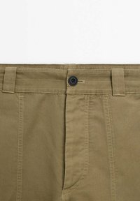 Gros plan sur la ceinture d’un pantalon vert olive avec un seul bouton noir, des passants de ceinture et des coutures visibles sur le tissu.