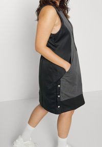 Femme en robe sans manches noire et grise mi-longue avec poches latérales et boutons-pression, portant des chaussettes blanches et des baskets, debout de profil.