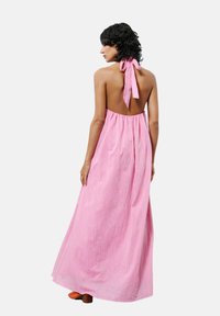 Roze, halter maxi-jurk zonder achterkant met een gestrikt bandje rond de hals, een vloeiende silhouet en lichte, gestructureerde stof. Model draagt oranje hakken.