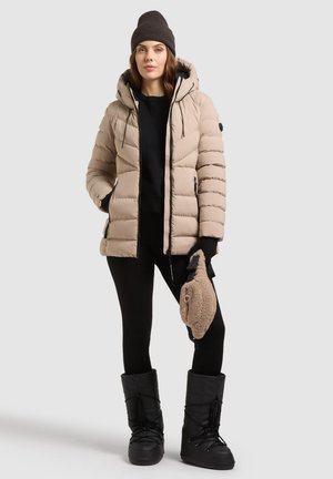 Une femme porte une doudoune beige à capuche, un bonnet noir, un legging et des bottes. Elle tient un petit sac beige en tissu sherpa, sur un fond uni.