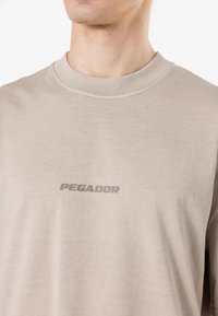 Bež majica s dolgimi rokavi iz mehkega bombaža z rebrastim izrezom. Na prsih je svetlo siva logotip "PEGADOR".