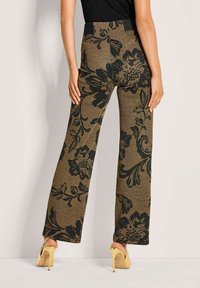 Vrouw in hoge taille, wijdvallende bruine broek met een groot zwart bloemenpatroon en gele hoge hakken, staande tegen een lichte muur.