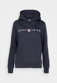 Granatowy hoodie wykonany z miękkiego materiału, z długimi rękawami, kapturem z troczkami oraz logo "GANT 1949" z detalem w kształcie tarczy.