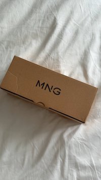 Obdĺžniková hnedá kartónová krabica s čiernym logom „MNG“ umiestneným uprostred vrchu, položená na pokrčenej bielej látke.