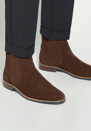 Bottines en daim marron avec semelles style bois portées avec un pantalon noir retroussé, présentées sur un fond clair.