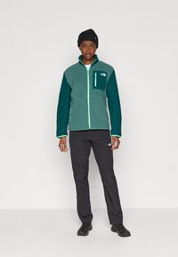 Flisjakke i mørk teal med lysegrønne detaljer, glidelåslomme, og svarte bukser. Modell iført svart beanie og svarte sko.