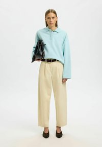 Hellblaues Polohemd mit langen Ärmeln und geknöpftem Kragen, kombiniert mit cremefarbenen Hosen mit weitem Bein und dunkelbraunen Schuhen. Hält eine schwarze Tasche.