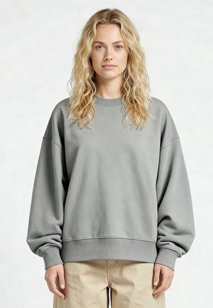 Jeune femme aux cheveux blonds ondulés portant un sweat-shirt gris surdimensionné et un pantalon beige, debout devant un fond clair uni.