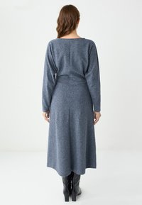 Robe en maille grise mi-mollet à manches longues vue de dos, avec une taille cintrée, une jupe légèrement évasée et des coutures minimales, associée à des bottes noires.