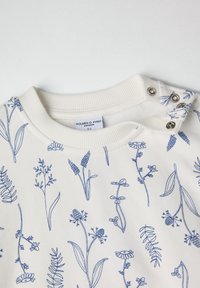 Huppari vaaleasta puuvillasta, jossa on sininen kukkakuosi. Pyöreä kaula-aukko, jossa ribattu viimeistely ja kaksi nepparia olkapäällä helppoa pukemista varten.