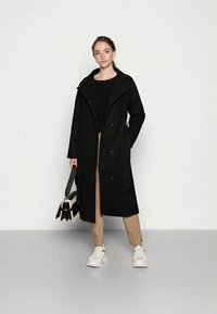 Giovane donna che indossa un lungo cappotto nero, pantaloni beige, sneakers bianche, mentre tiene una borsa in bianco e nero, in piedi contro uno sfondo chiaro e semplice.
