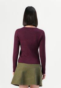 Femme aux cheveux noirs raides portant un haut violet à manches longues côtelé et une jupe patineuse vert olive, tournée dos à un fond uni.