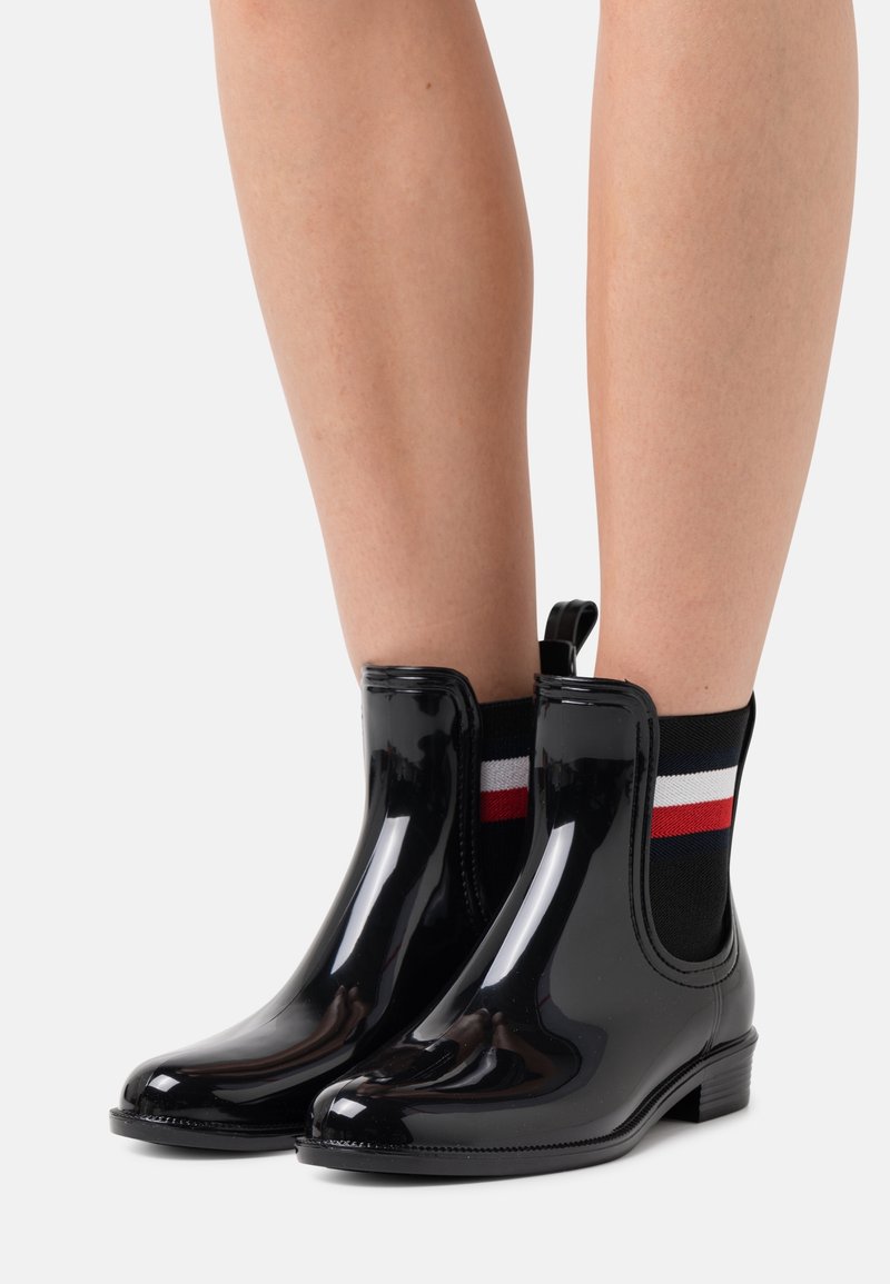 Bottes noires en caoutchouc à la cheville avec une finition brillante, des panneaux latéraux élastiques et une petite languette ; associées à des chaussettes noires avec des rayures rouges et blanches.