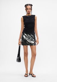 Mulher vestindo uma blusa preta sem mangas, saia mini de lantejoulas prateadas, sandálias de salto pretas, segurando uma bolsa preta contra um fundo simples.