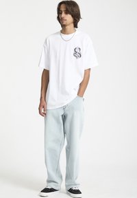 T-shirt en coton blanc avec un motif de serpent graphique sur la poitrine gauche, associé à un jean en denim bleu clair et des baskets noires.